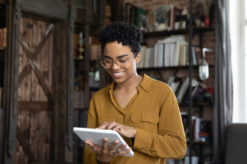 Smiling millennial African American woman in glasses using digital computer tablet applications,...