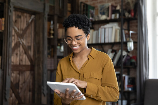 Smiling millennial African American woman in glasses using digital computer tablet applications, enjoying playing games, shopping in internet store, communicating in social network, web surfing.