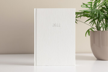 White book mockup, diary for 2022 and green plant in pot on white table against background of beige wall. Front view. Place for text, copy space, mockup