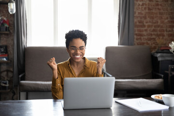 Happy laughing young African American woman in glasses looking at computer screen, clenching fists...