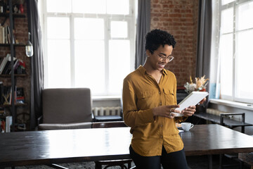 Happy young African American woman in eyeglasses using digital computer tablet, leaning on table in modern home office, enjoying communicating distantly, checking email, shopping online, copy space. - obrazy, fototapety, plakaty
