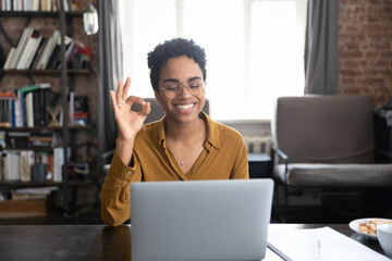 Happy young African American woman in glasses showing okey gesture, looking at computer screen,...