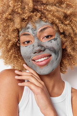 Vertical shot of gentle smiling woman touches jawline gently applies clay nourishing mask for skin...