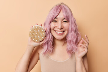 Happy woman with eastern appearance holds brush for massage tries to reduce cellulite has pink dyed hair smiles toothily enjoys body care treatment isolated over beige background feels joyful