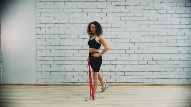 Workout in sports studio - young woman with curly hair training her hand with an elastic band by stepping on it and pulling motions