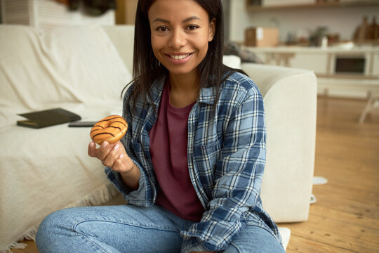 Close-up image of charming 20-years-old female in blue plaid shirt upon pink t-shirt and jeans holding desired glazed and decorated doughnut, waiting with anticipation to eat it, looking excited