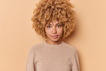 Portrait of serious confident sassy good looking woman with bushy curly hair looks directly at camera stands indoor against beige background wears casual jumper. Human face expressions concept © WHstudio Leushin N