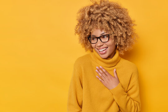 Positive Sincere Curly Woman Keeps Hand On Chest Looks Away Happily Smiles Broadly Laughs At Something Funny Wears Spectacles And Casual Jumper Isolated Over Yellow Background. Emotions Concept