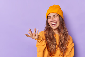 Obraz na płótnie Canvas Happy teenage girl raises hand looks joyfully at something funny wears hat and jumper poses against purple background with blank space for your advertising content. Positive human emotions concept