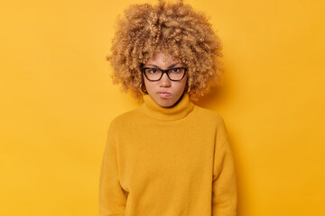 Fototapeta na wymiar Horizontal shot of outraged curly haired woman frowns face looks angrily being irritated with someone wears transparent glasses and casual sweater isolated over yellow background. Negative emotions
