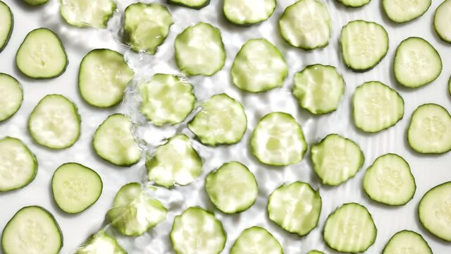Wide shot of poured water making splashes and ripples over cucumber slices arranged in rows on white background | makeup background, cleanser commercial