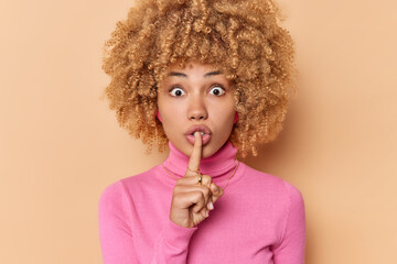 Surprised curly haired woman presses index finger over lips asks to keep promise has shocked expression makes silence gesture dressed in casual pink turtleneck isolated over beige background