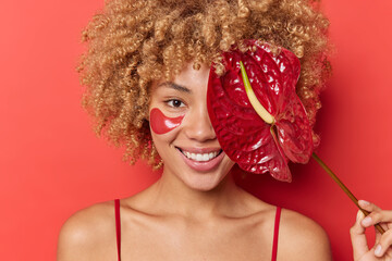 Portrait of happy woman covers face with red calla lily applies beauty patches for skin refreshing stands bare shoulders smiles gladully isolated over bright background. Wellness and natural cosmetics