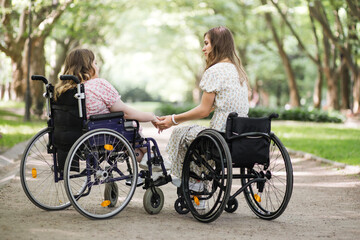 Back view of two young women in summer dressed holding hands while sitting in wheelchairs among green park. People who live with disability supporting each other.