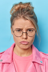 Vertical shot of dissatisfied sulking woman has angry offended expression has discontent expression wears round spectacles pink jacket isolated over blue background. Negative human emotions.