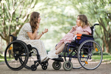 Side view of two happy women in wheelchairs talking and smiling outdoors. Background of green summer park. Concept of people with chronic health condition.