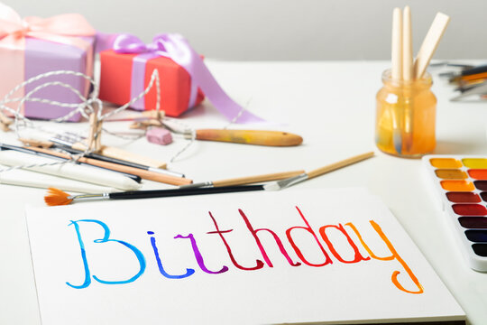 The word "Birthday" is painted on a white sheet of paper in multicolored watercolors. Preparing for the holiday with your own hands. Do it yourself.
