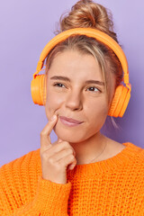 Thoughtful dreamy woman keeps index finger near corner of lips being deep in thoughts while listens lyrics songs in headphones dressed casually isolated over purple background. Let me think.