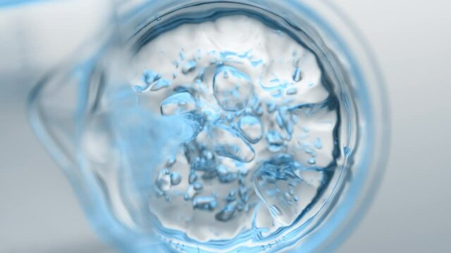 Light blue liquid pouring into beaker with water from flask creates different sized bursting bubbles and ripples on its surface on grey background | Abstract skin cleanser mixing concept
