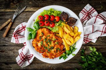Breaded fried pork chop with French fries and vegetables on wooden table
