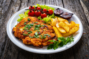 Breaded fried pork chop with French fries and vegetables on wooden table
