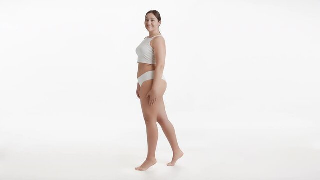 Horizontal long shot of young pretty Caucasian plus size woman in white underwear stands on tiptoes and puts her hand on her waist smiling wide to the camera on white background | Body care concept