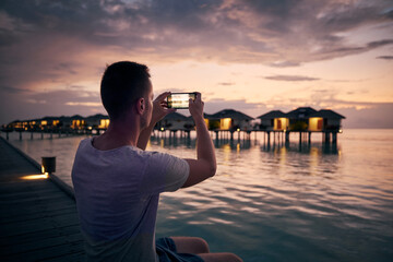 Young man with mobile phone photographing seascape with water bungalows at beautiful sunset in Maldives..