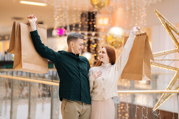 Portrait of happy beautiful young couple in love holding shopping bags standing in mall and looking at each other. Joyful handsome man and attractive woman purchasing together at store center.