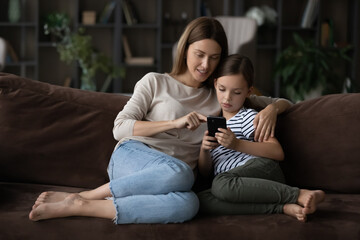Spend time together. Caring millennial female teach preteen daughter younger sister child use mobile telephone app. Bonding young mother and tween kid girl cuddle on cozy couch make video call on cell