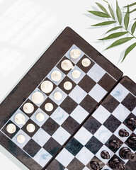 Chess game. Chess composition with a chess board with placed pieces and palm leaves on a blue background. Chess competition. Flat lay