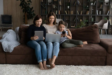 Family online. Three women relatives of diverse age sit on sofa at living room use digital gadgets. Old grandmother young adult mother preteen schoolgirl daughter grandchild hold laptop touchpad phone