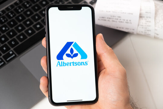 Albertsons application of supermarket and grocery store in a smartphone. A man is holding a smartphone with app for shopping and laptop on the background. November 2021, San Francisco, USA