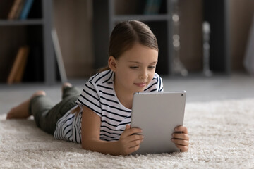 Kid tablet user. Little preteen age girl lie on fluffy carpet hold digital pad pc look at screen...