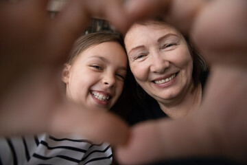 With love and care. Smiling old age granny tween girl grandkid look at camera through tender heart of joined fingers. Aged woman custodian shoot affectionate selfie portrait with preteen foster child - Powered by Adobe