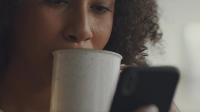 Modern morning habits. Close up shot of young african american woman drinking hot coffee and reading news on smartphone