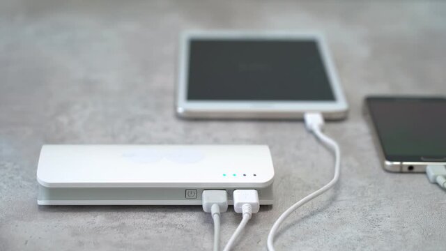Male hand turns on powerbank on background of mobile phone and digital tablet. External battery charges electronic devices. Flashing indicators