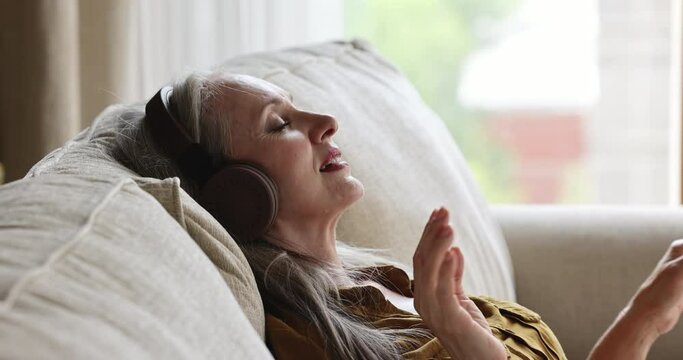 Beautiful aged female music lover rest on couch in modern headset listen to classical melody compositions enjoy perfect quality sound. Carefree mature lady having pleasure hearing songs keeping rhythm