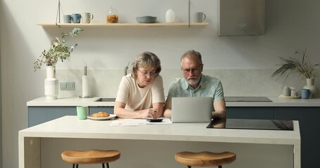 Serious older family couple sit at home office in cozy kitchen manage domestic finances together use laptop. Busy senior spouses retirees discuss taxes bills pay utilities online via web bank service
