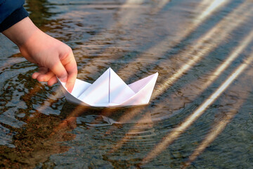 A child lowers a paper boat into the water . A close - up of a child 's hand holding a boat in the water . Happy childhood . Playing outside .