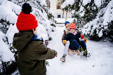Fototapeta na wymiar Two children and their laughing mother enjoying sleigh ride. Children play outdoors in snow. Kids sled in the in winter snowy city park arround snowy fir trees. Outdoor fun for family vacation.