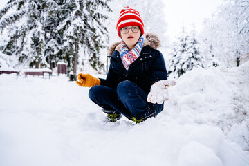 Cute handsome boy down syndrome in eyeglasses and bright wear looking at camera playing with snow outdoor in frosty snowy winter park.