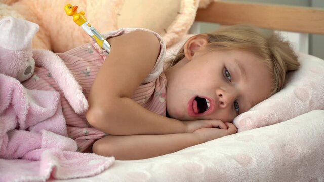 Sick little girl lies in bed at home with thermometer and coughs. Temperature measurement in illness. Treatment for flu and cold