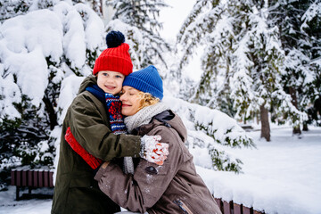 Fototapeta na wymiar Happy family moments, wnter games concept. Side view of happy mother carrying and hugging cute little son together while standing against snowy trees in forest or park.