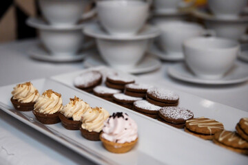 Babeczki i ciasteczka zaserwowane koło filiżanek przygotowanych do parzenia kawy i herbaty w restauracyjnej sali konferencyjnej. - obrazy, fototapety, plakaty