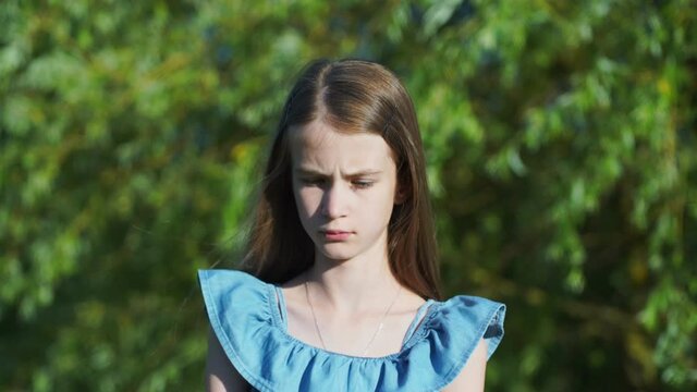 Unhappy teenage girl with loose fair hair in blue blouse looks into camera against blurry green trees in sunny summer park