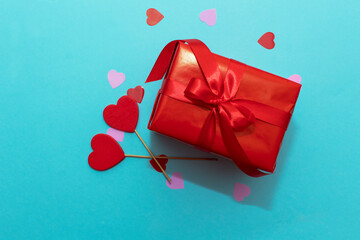 Red box with red ribbon on a blue paper background. Valentine's Day gift with confetti and decor