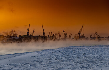 The construction of nuclear icebreakers at magic sunset, cranes of of the Baltic shipyard in a frosty winter day, steam over the Neva river, smooth surface of the river, sky of orange color