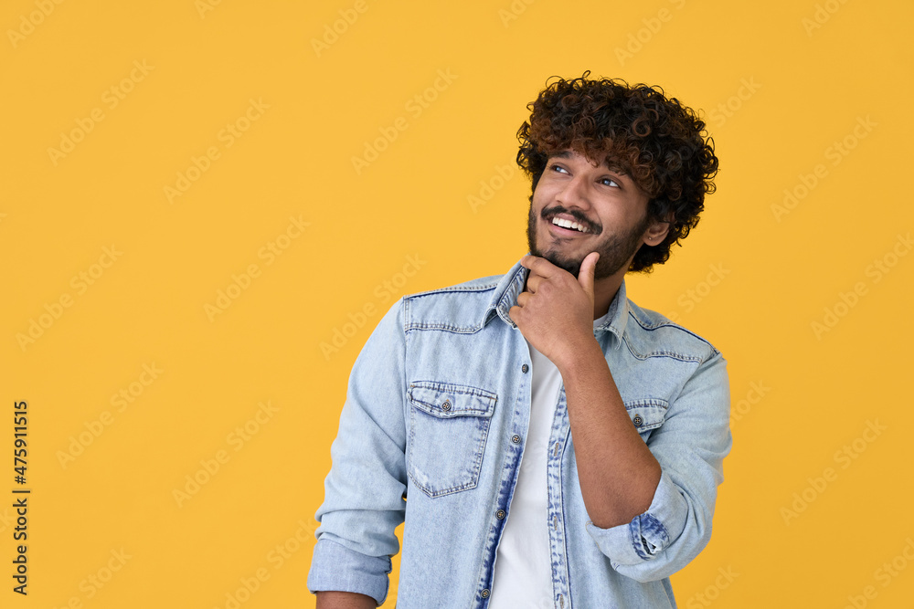 Wall mural Smiling curious young indian man holding hand on chin looking interested aside at copy space isolated on yellow background thinking of shopping opportunities, planning purchase or dreaming concept.