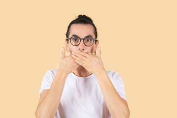 handsome latin man with glasses to see, covers his mouth face of surprise or fright, beige background