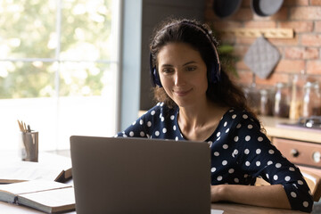 Pleasant happy beautiful young hispanic woman wearing headphones, watching interesting online lecture on computer, improving knowledge distantly on internet courses, communicating with tutor remotely.